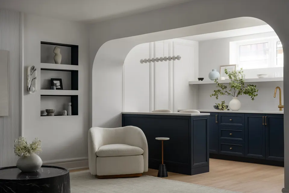 Basement lounge framed by a plaster arch: a navy shaker wet bar with a brass tap, open shelving, a cream bouclé armchair and display niches