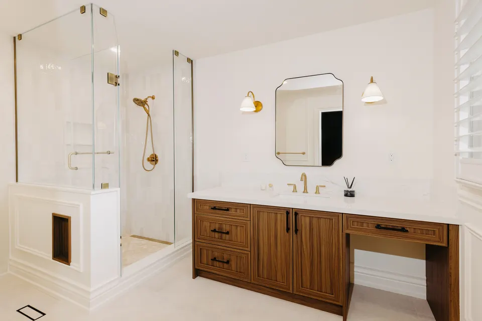 Bright ensuite: a walnut shaker vanity and makeup area under a scalloped mirror, a glass shower with a half wall, white wall mouldings