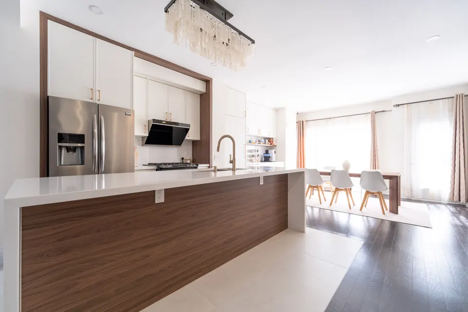 A long waterfall island in walnut and white quartz running toward the dining area, a crystal chandelier above