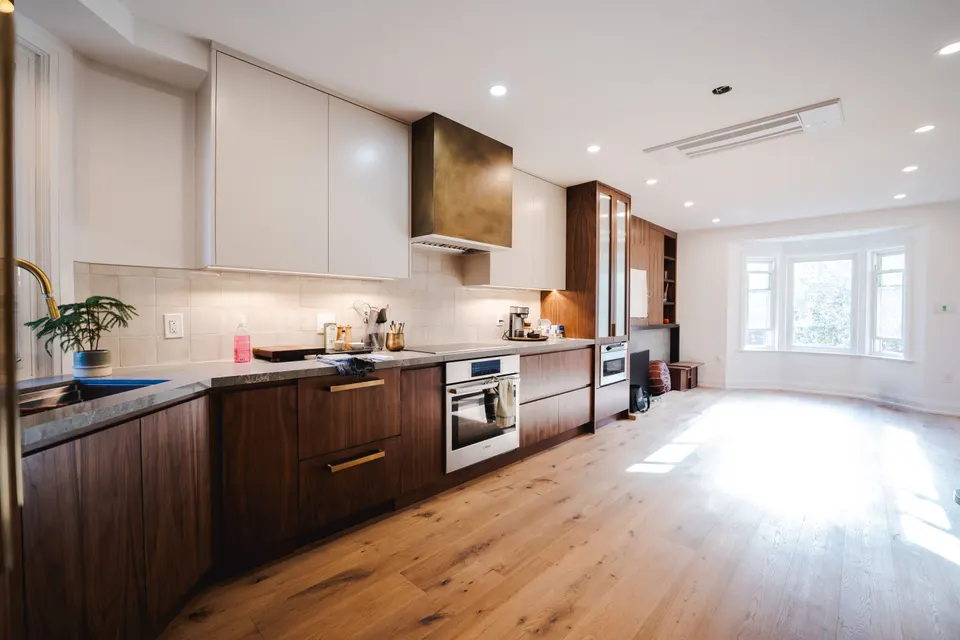 Open-plan second floor: walnut lower cabinets and a bronze range hood run toward a bay window, wide-plank oak floor in afternoon light