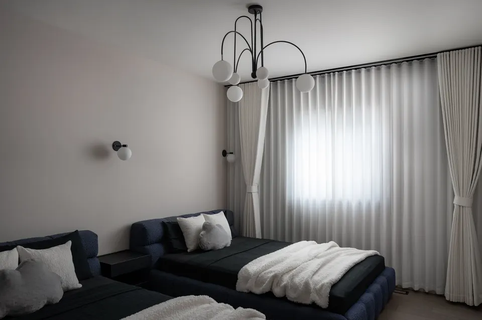 Bedroom with two low navy beds under white throws, sheer curtains drawn across a tall window and a black arched chandelier hung with glass globes
