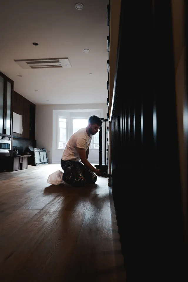A tradesman kneeling at the foot of a new black stair in a long, sunlit room with oak floors