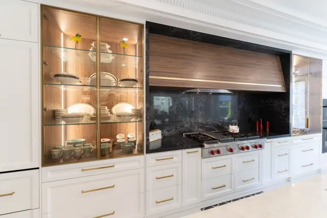 White kitchen with lit glass-front cabinets either side of a walnut range hood and dark stone backsplash