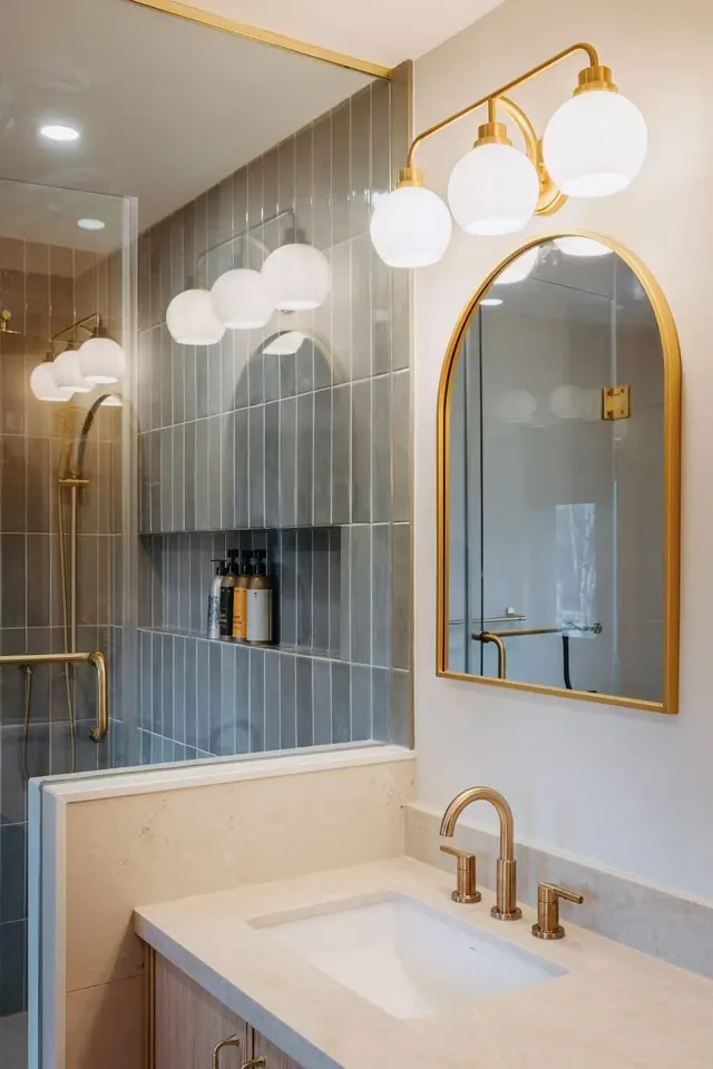 Arched brass mirror and globe sconces over a vanity, sage tile shower beside