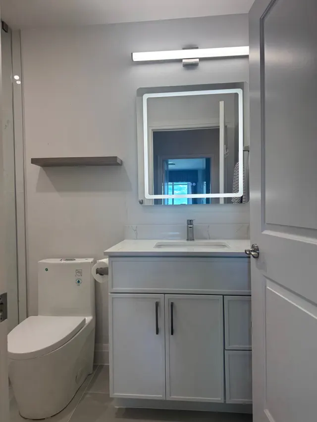 Grey condo vanity with a white top under a lit mirror and a light bar, beside the toilet