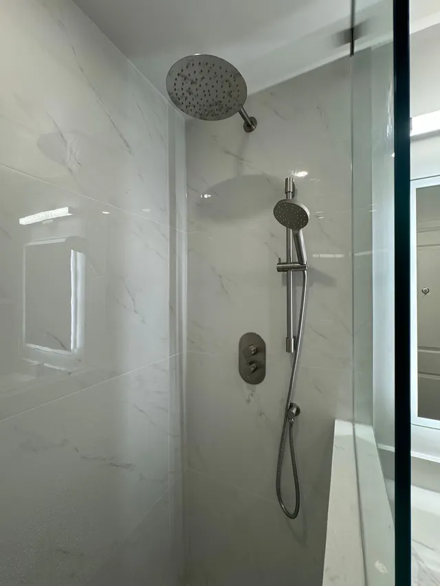 A rain shower head and a hand shower on white marble-look tile behind frameless glass